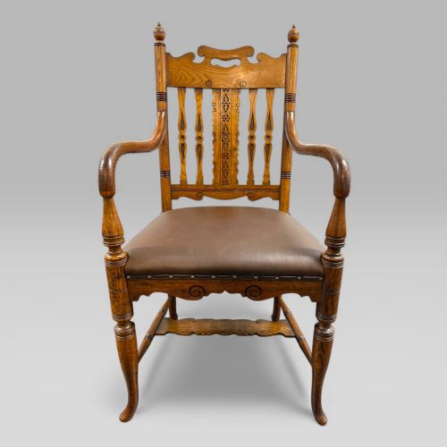 fig 1. A Victorian golden oak armchair featuring a carved backrest, curved arms, brown leather seat with brass studs, and turned legs.