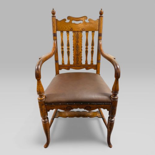 fig 3. A Victorian golden oak armchair featuring a carved backrest, curved arms, brown leather seat with brass studs, and turned legs.