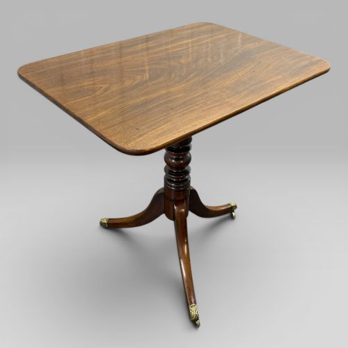 fig 1. A Regency period mahogany tilt-top occasional table featuring a rectangular surface, turned central pedestal, and tripod legs ending in brass castors.