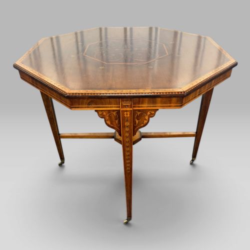fig 3. An octagonal Edwardian mahogany centre table featuring floral marquetry inlays, satinwood crossbanded borders, tapered legs, an X-stretcher, and brass castors.