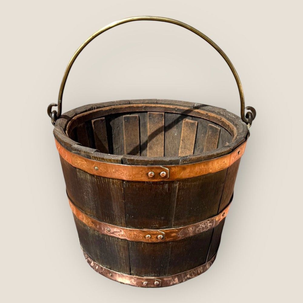 An antique coopered oak bucket featuring multiple riveted copper bands and a curved metal handle against a neutral background.