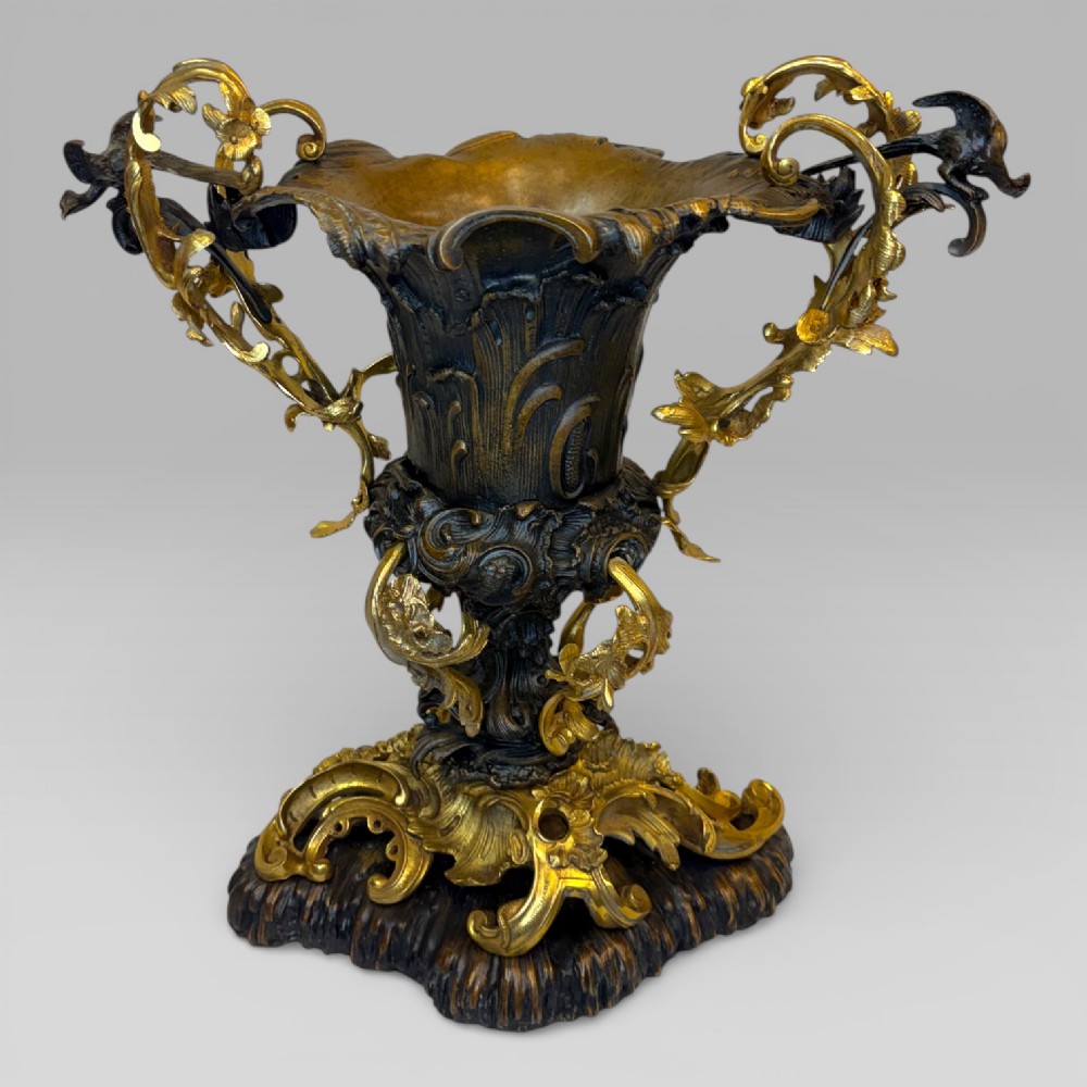 An ornate gilt and patinated bronze centrepiece urn featuring twin scrolled foliate handles, a scalloped rim, and a sculpted rocaille base.