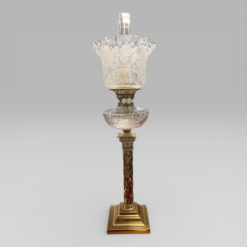 An antique Victorian oil lamp featuring a Corinthian style brass column base, clear glass reservoir, and an etched glass tulip shade with a frilled top rim.