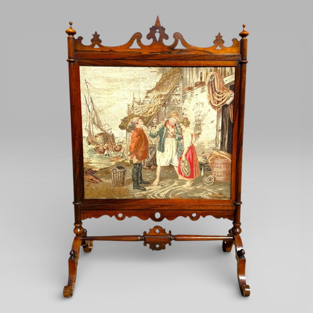 A Victorian rosewood cheval fire screen featuring a carved wooden frame with finials and an embroidered textile panel depicting figures near a coastal cottage.