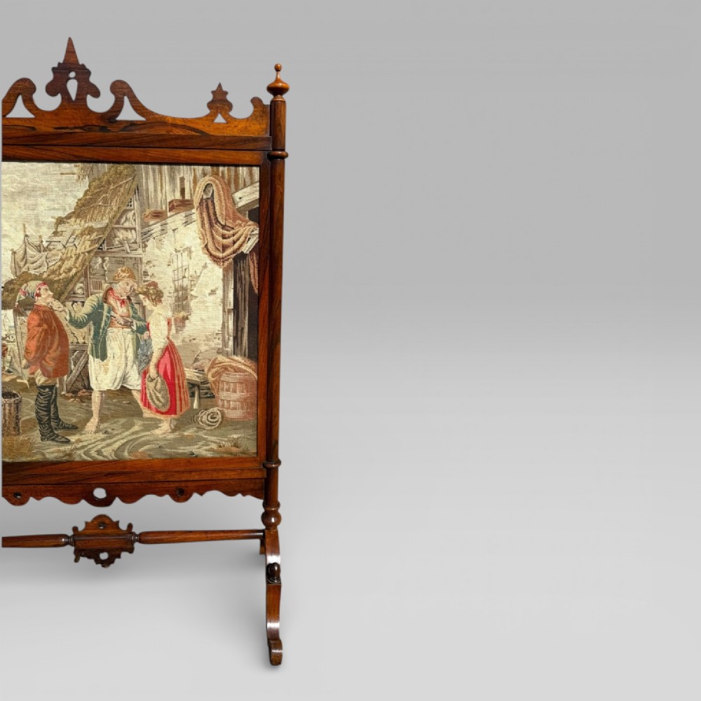 A Victorian rosewood cheval fire screen featuring a carved wooden frame with finials and an embroidered textile panel depicting figures near a coastal cottage. View V