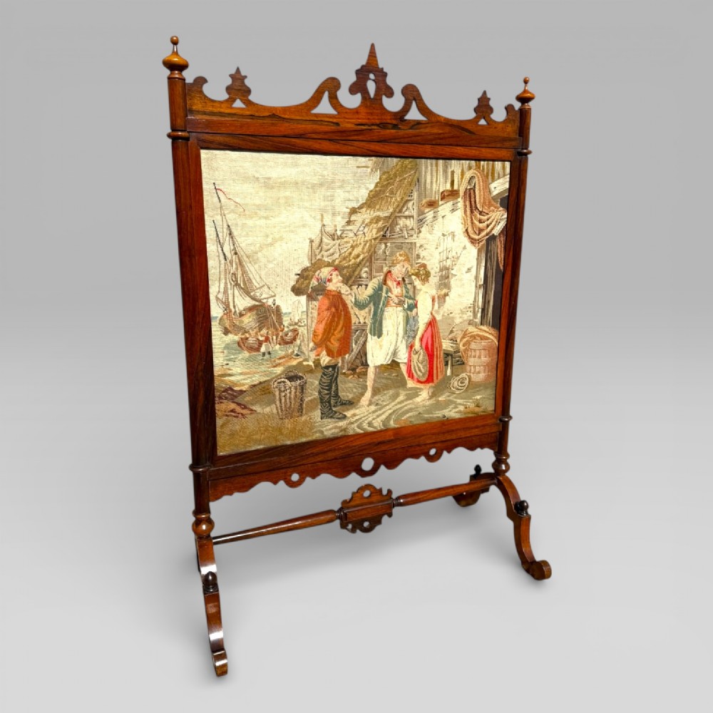A Victorian rosewood cheval fire screen featuring a carved wooden frame with finials and an embroidered textile panel depicting figures near a coastal cottage. View VI