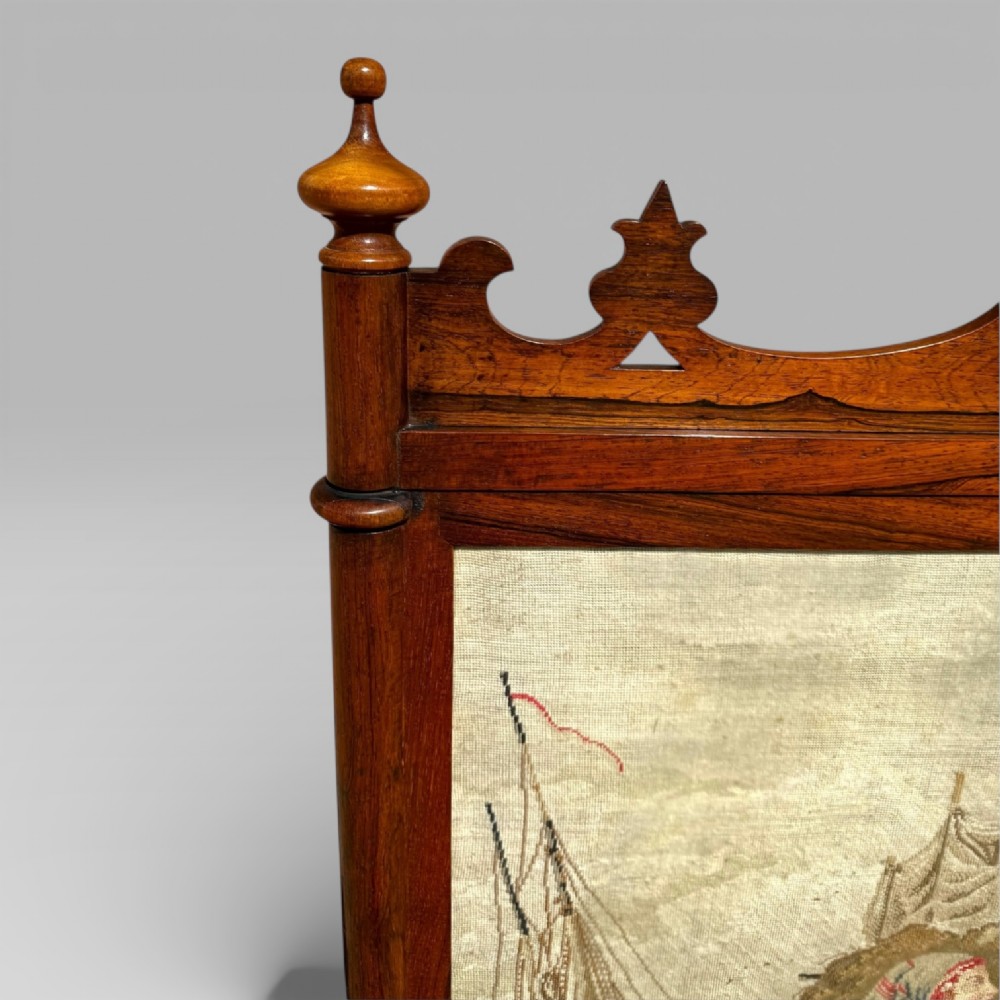 A Victorian rosewood cheval fire screen featuring a carved wooden frame with finials and an embroidered textile panel depicting figures near a coastal cottage. View VIII