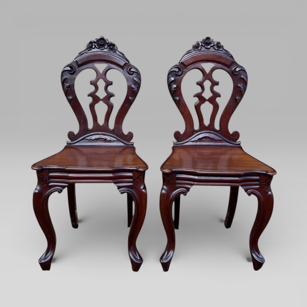 A matching pair of antique Victorian mahogany hall chairs featuring carved pierced backrests, solid wooden seats, and cabriole front legs.