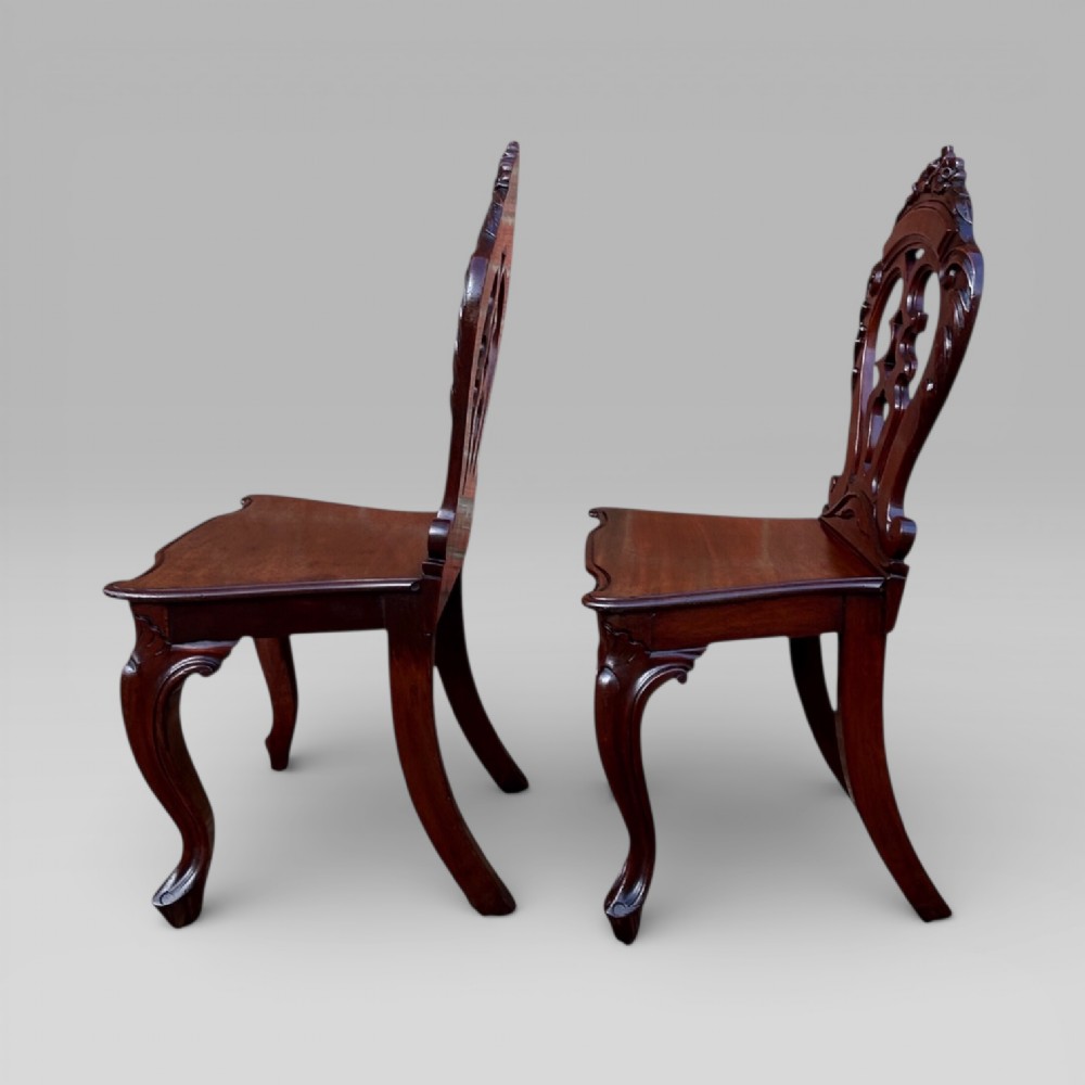 A matching pair of antique Victorian mahogany hall chairs featuring carved pierced backrests, solid wooden seats, and cabriole front legs. View V