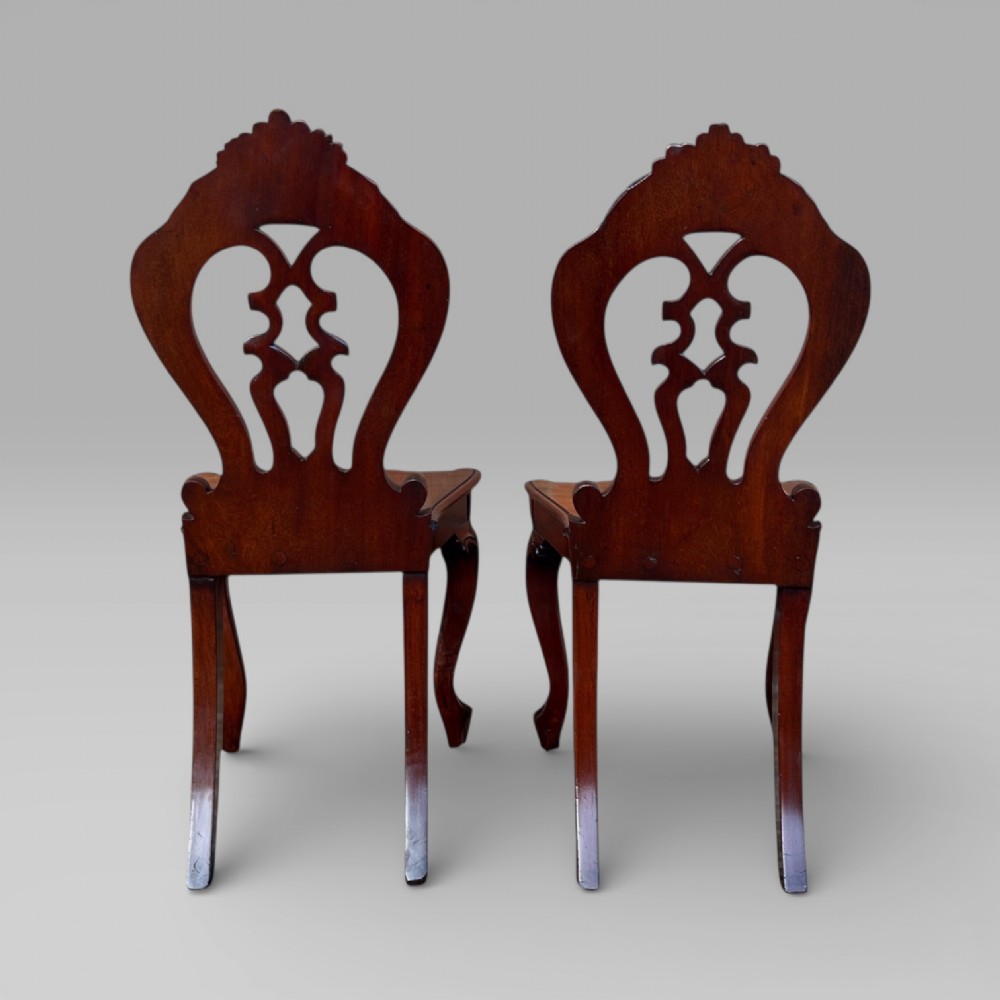 A matching pair of antique Victorian mahogany hall chairs featuring carved pierced backrests, solid wooden seats, and cabriole front legs. View VI