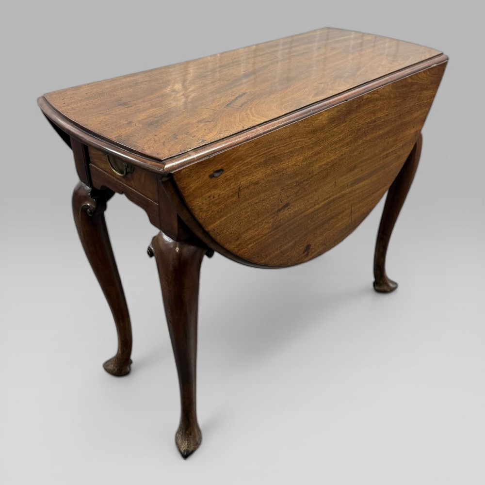 An 18th-century George II mahogany drop-leaf table with hinged oval flaps, cabriole legs terminating in pad feet, and a side drawer with a brass handle.