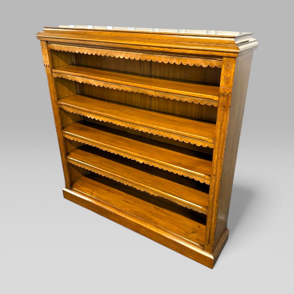 A late Victorian open bookcase crafted from golden oak, featuring scalloped edging along each shelf and subtle geometric carvings on the pilasters.