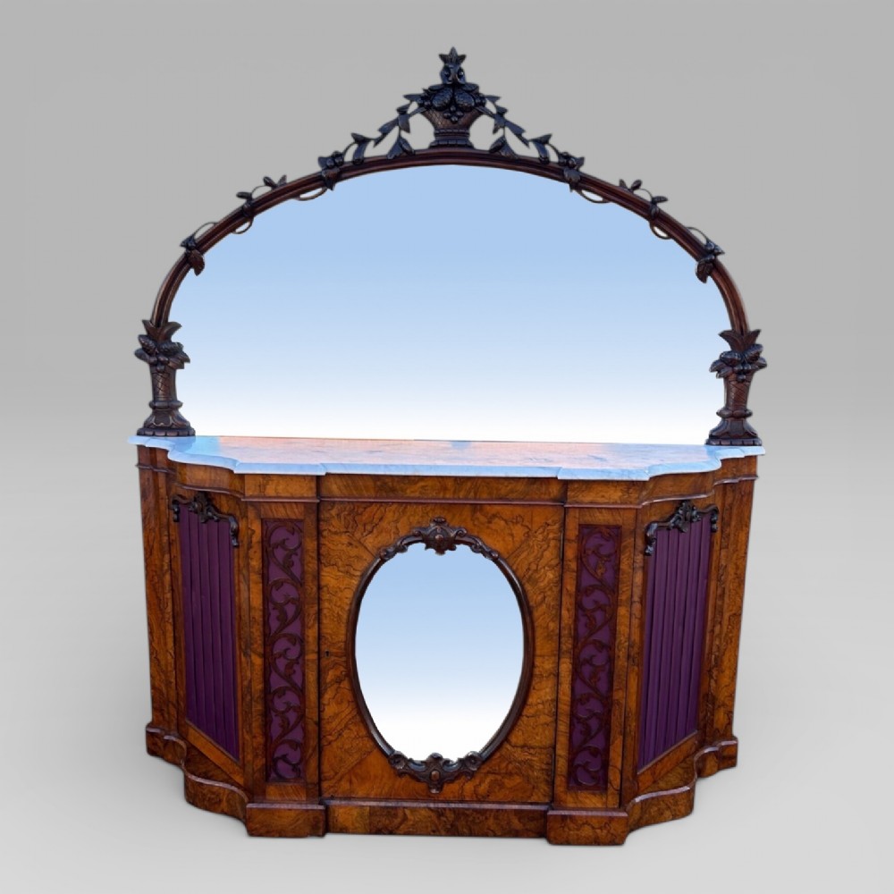 A Victorian walnut credenza featuring a breakfront white marble top, an arched crest-carved mirror frame, and base panels detailed with an oval mirror and fabric behind fretwork.
