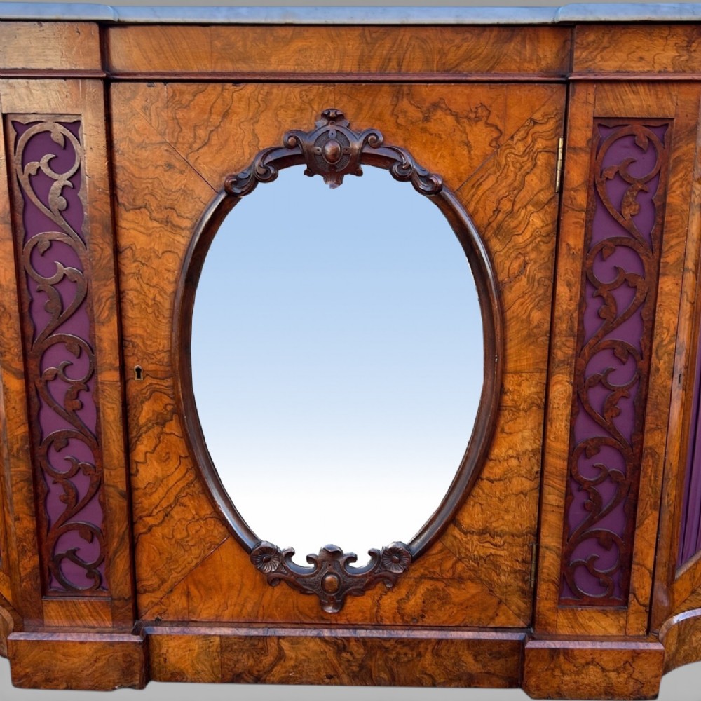 A Victorian walnut credenza featuring a breakfront white marble top, an arched crest-carved mirror frame, and base panels detailed with an oval mirror and fabric behind fretwork. View V