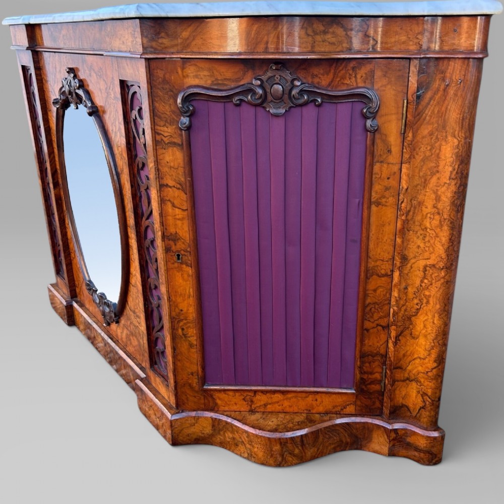 A Victorian walnut credenza featuring a breakfront white marble top, an arched crest-carved mirror frame, and base panels detailed with an oval mirror and fabric behind fretwork. View VI