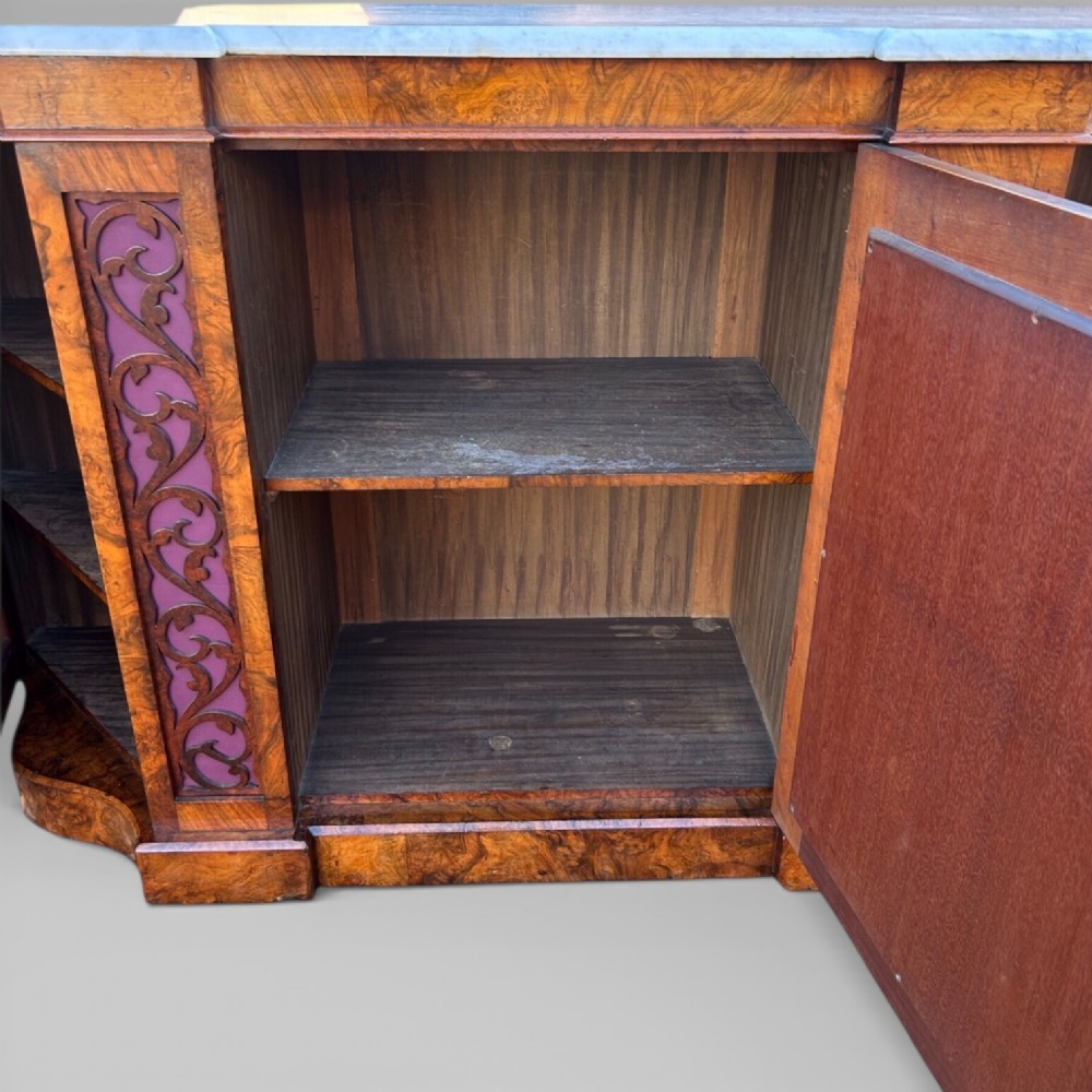 A Victorian walnut credenza featuring a breakfront white marble top, an arched crest-carved mirror frame, and base panels detailed with an oval mirror and fabric behind fretwork. View VII