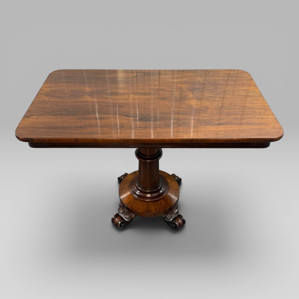 An antique William IV rosewood centre table featuring a rectangular top with rounded corners resting on a central turned pedestal and carved feet.