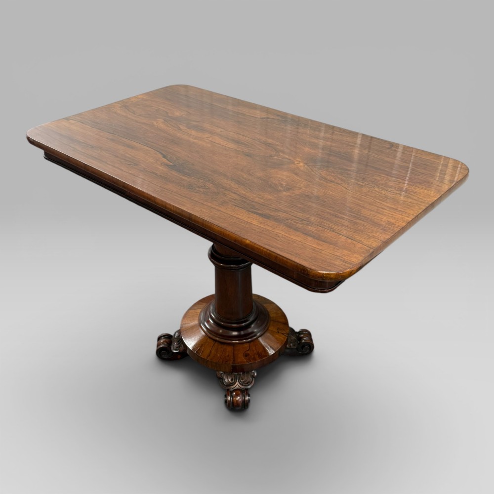 An antique William IV rosewood centre table featuring a rectangular top with rounded corners resting on a central turned pedestal and carved feet. View II