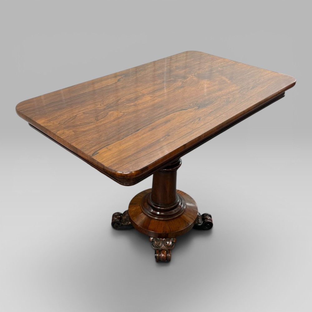 An antique William IV rosewood centre table featuring a rectangular top with rounded corners resting on a central turned pedestal and carved feet. View III