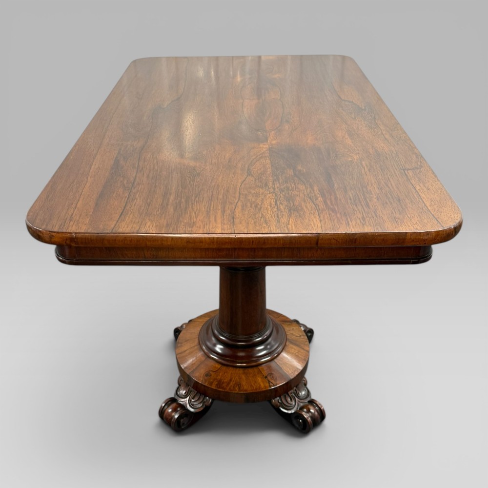 An antique William IV rosewood centre table featuring a rectangular top with rounded corners resting on a central turned pedestal and carved feet. View V