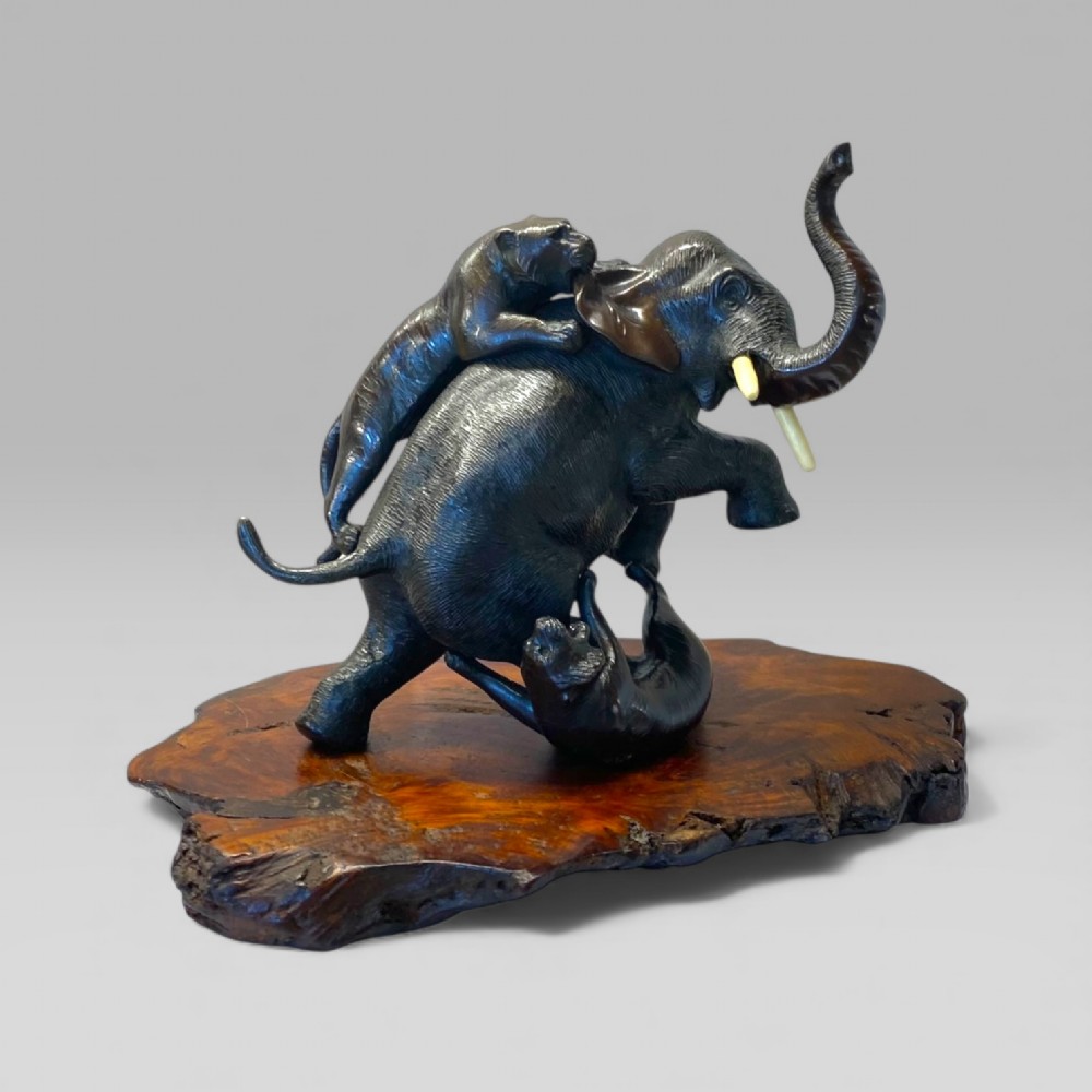 A dark patinated bronze sculpture depicting an elephant defending itself against two tigers, mounted on a polished natural edge wooden base. View II