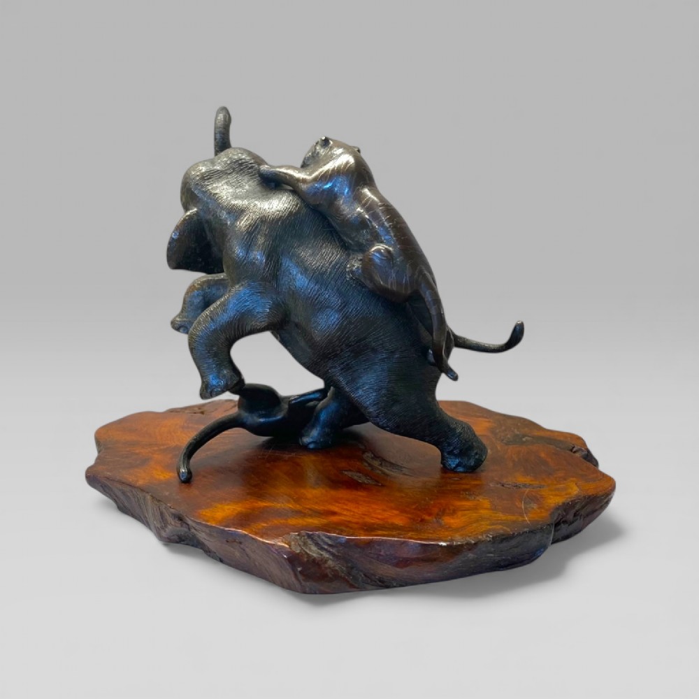 A dark patinated bronze sculpture depicting an elephant defending itself against two tigers, mounted on a polished natural edge wooden base. View III