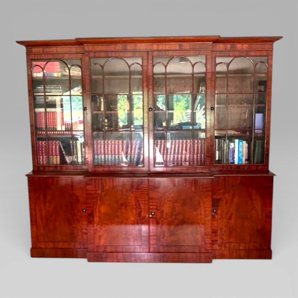 A large William IV flame mahogany breakfront library bookcase featuring four glazed upper doors with arched astragal glazing and four panelled cupboard doors beneath.
