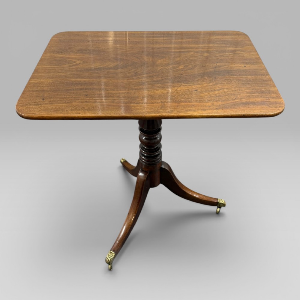 A Regency mahogany tilt-top table featuring a rectangular surface, turned central pedestal column, and tripod splayed legs with brass lion paw castors.