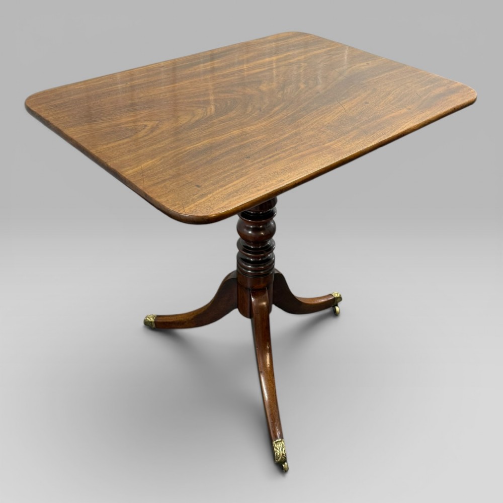 A Regency mahogany tilt-top table featuring a rectangular surface, turned central pedestal column, and tripod splayed legs with brass lion paw castors. View II