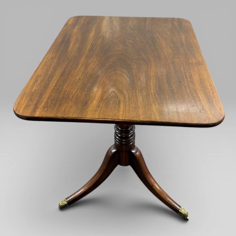 A Regency mahogany tilt-top table featuring a rectangular surface, turned central pedestal column, and tripod splayed legs with brass lion paw castors. View IV
