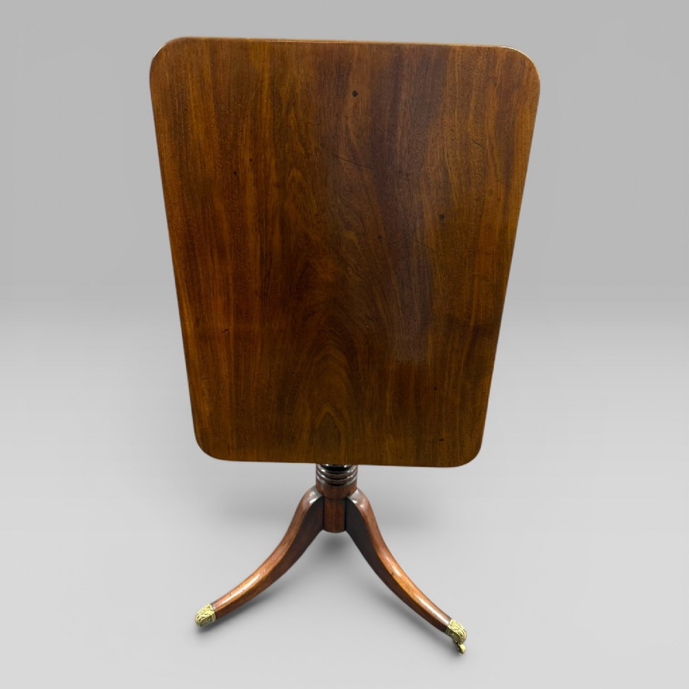 A Regency mahogany tilt-top table featuring a rectangular surface, turned central pedestal column, and tripod splayed legs with brass lion paw castors. View V