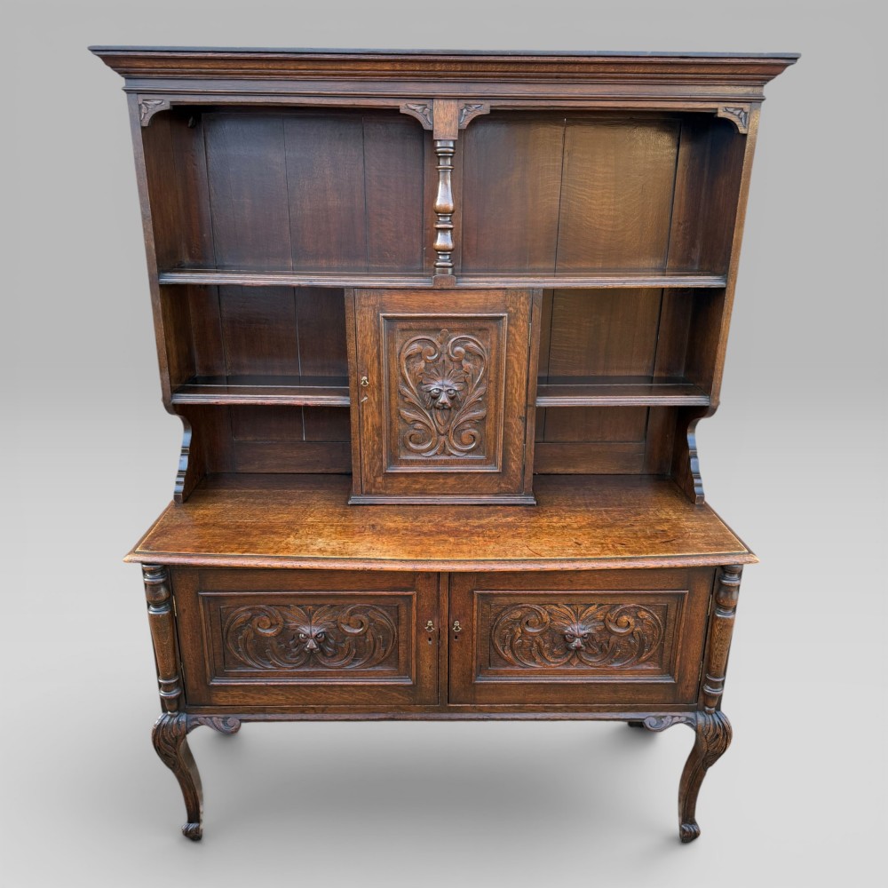 A late Victorian dark oak dresser with carved Green Man foliate motifs on the cupboard doors, open display shelving, and cabriole legs.