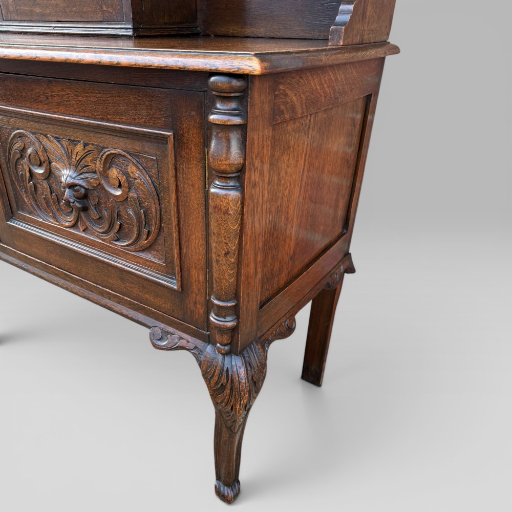 A late Victorian dark oak dresser with carved Green Man foliate motifs on the cupboard doors, open display shelving, and cabriole legs. View V