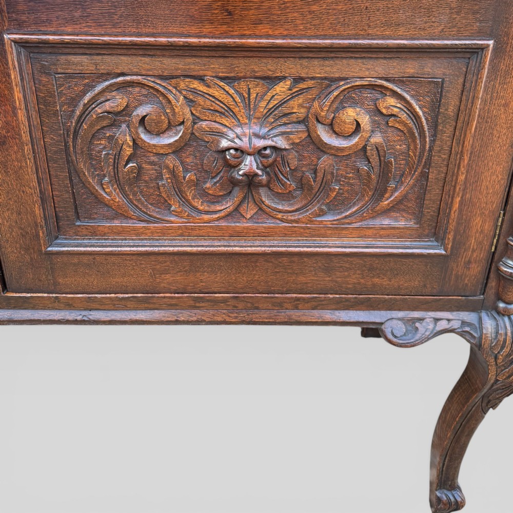 A late Victorian dark oak dresser with carved Green Man foliate motifs on the cupboard doors, open display shelving, and cabriole legs. View VI