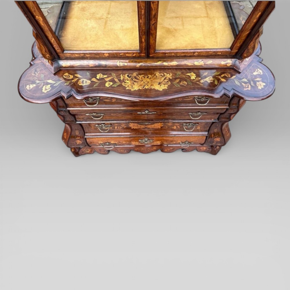 An antique walnut display cabinet on a bombe chest of drawers featuring floral marquetry inlay, glazed doors, and brass handles. View X