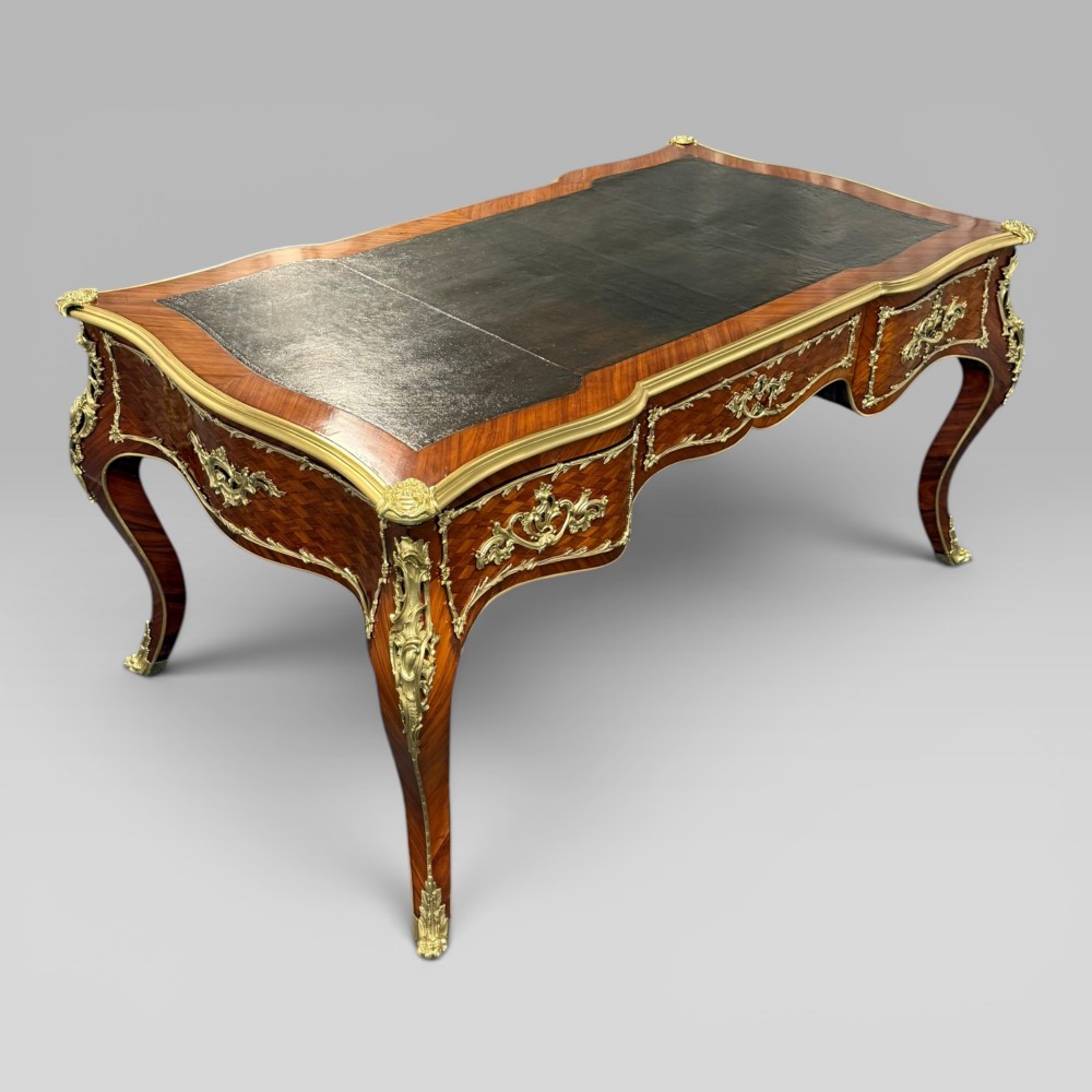 A Louis XV style kingwood parquetry bureau plat featuring a dark leather writing surface, cabriole legs, and gilt-bronze ormolu mounts.