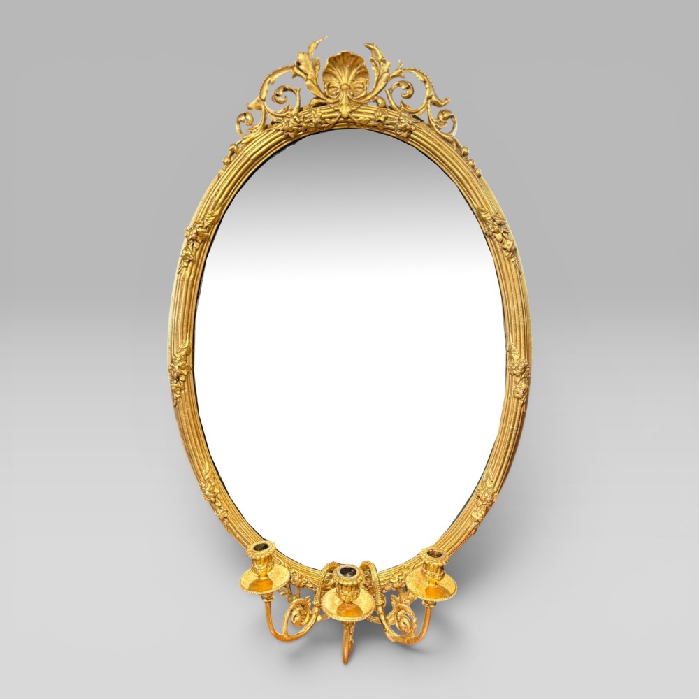 An oval giltwood mirror framed with floral carving and a top shell crest, featuring three candle sconces mounted at the base.