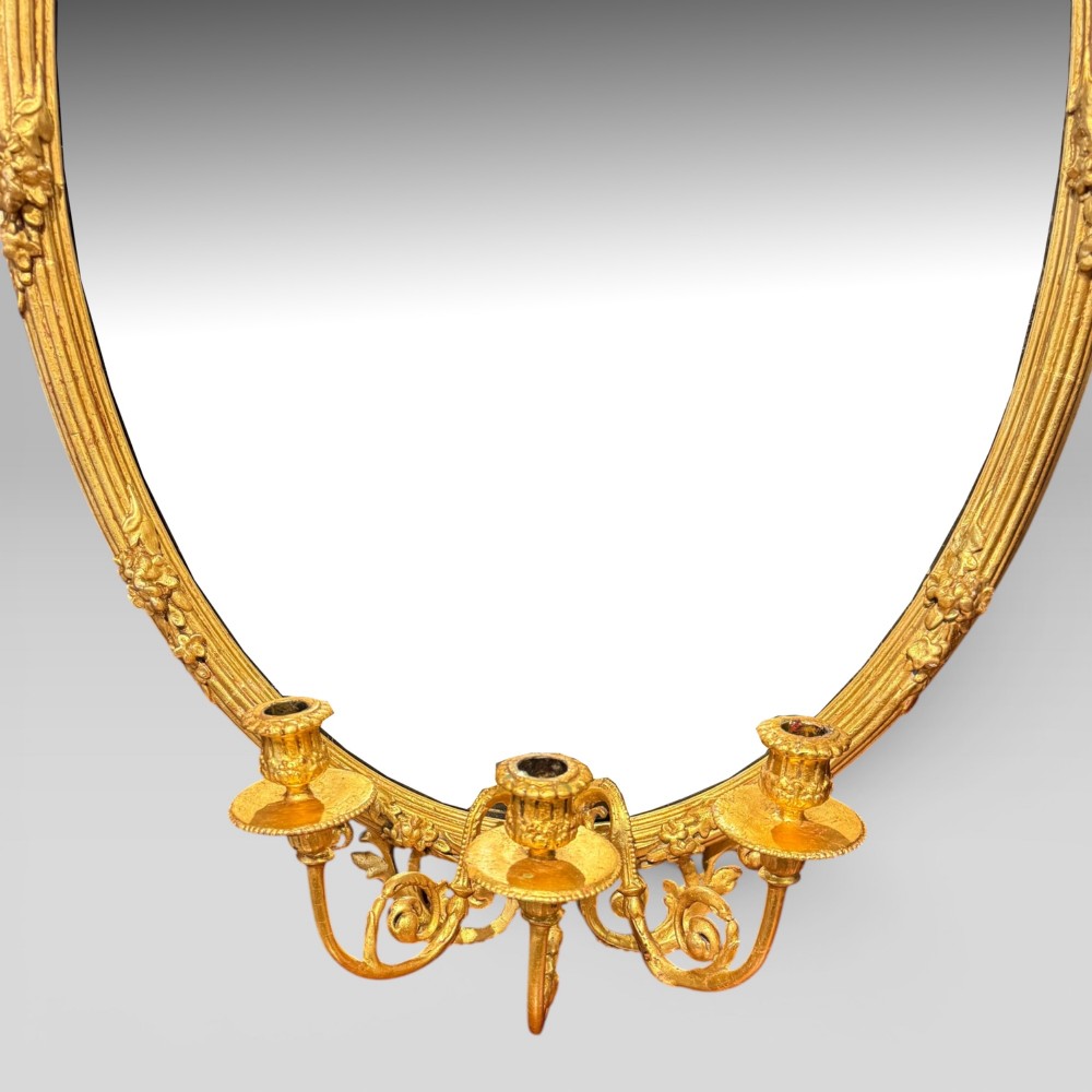 An oval giltwood mirror framed with floral carving and a top shell crest, featuring three candle sconces mounted at the base. View V