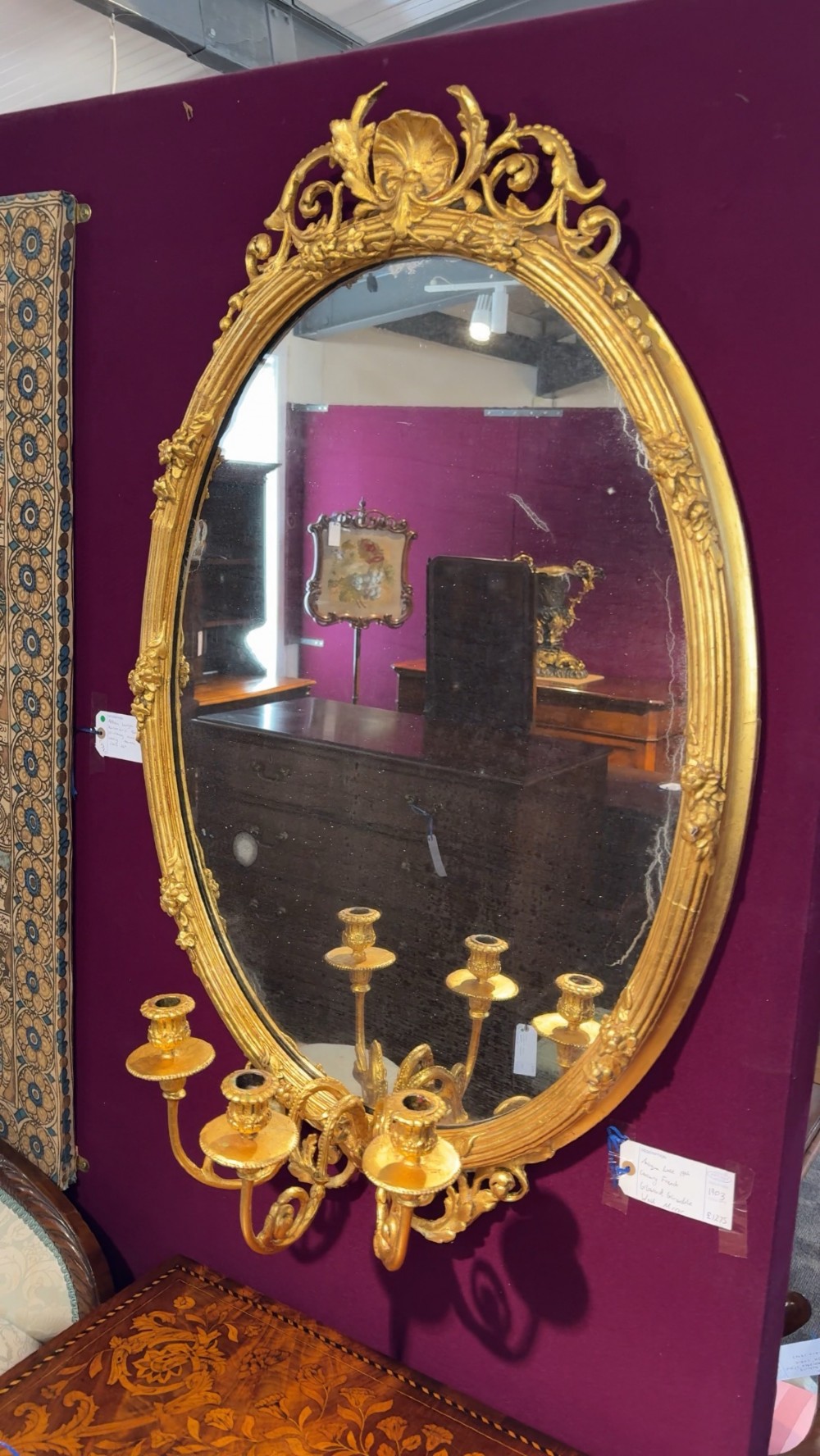 An oval giltwood mirror framed with floral carving and a top shell crest, featuring three candle sconces mounted at the base. View VI
