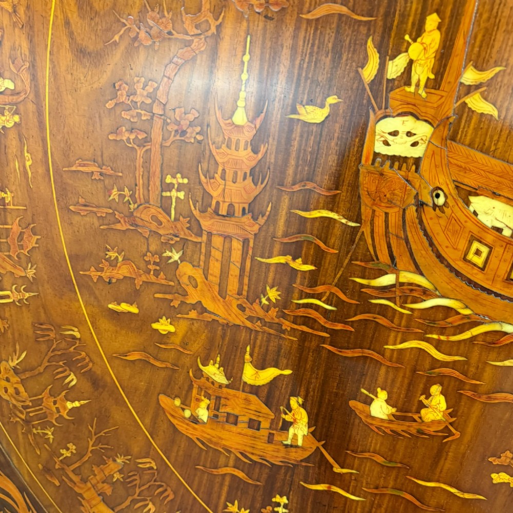Circular rosewood centre table featuring detailed marquetry depictions of ships, pagodas, and floral motifs resting on a square inlaid pedestal base. View X
