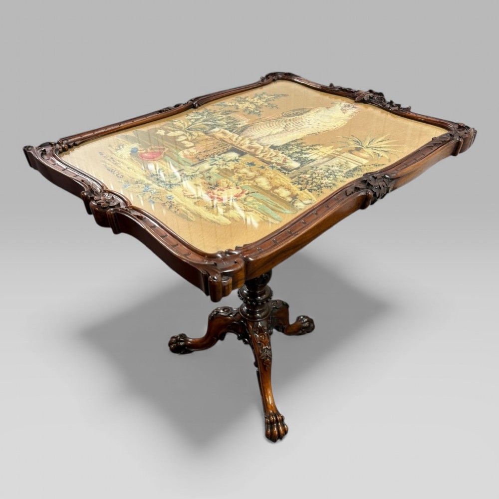 A William IV rosewood tilt-top tripod table featuring a shaped, carved frame housing a glazed needlepoint panel above a carved column and splayed tripod legs.