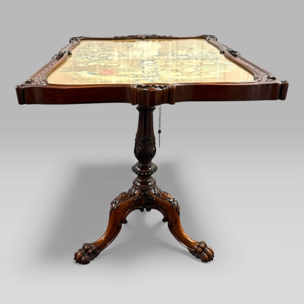 A William IV rosewood tilt-top tripod table featuring a shaped, carved frame housing a glazed needlepoint panel above a carved column and splayed tripod legs. View V