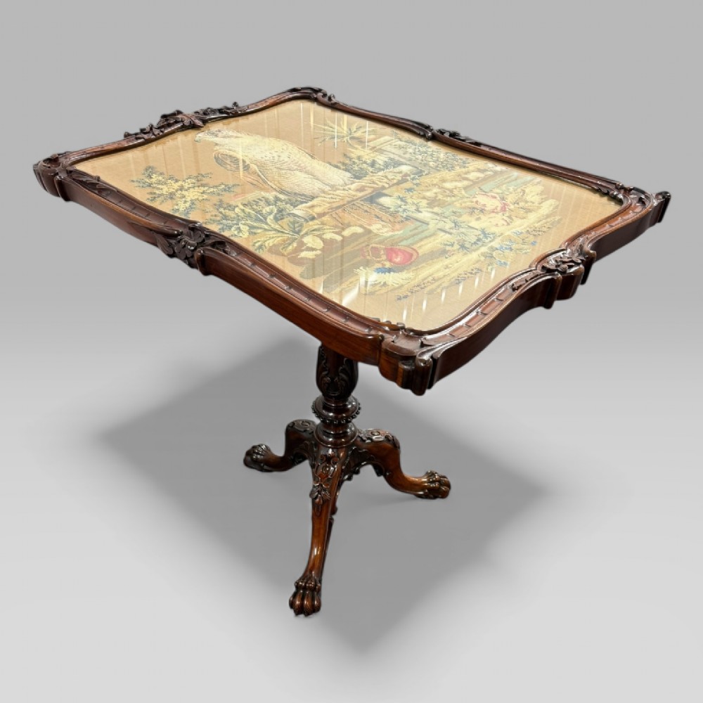 A William IV rosewood tilt-top tripod table featuring a shaped, carved frame housing a glazed needlepoint panel above a carved column and splayed tripod legs. View VI