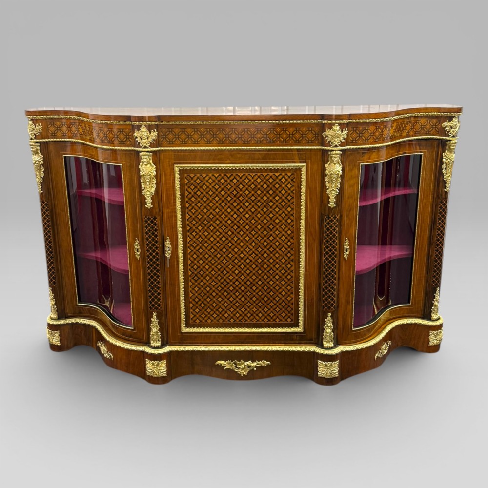 A Victorian walnut serpentine credenza featuring geometric marquetry, gilt ormolu mounts, a central panel door, and two curved glass side doors revealing lined display shelves.