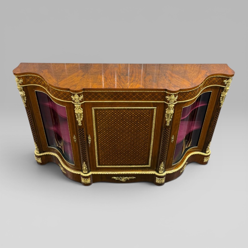 A Victorian walnut serpentine credenza featuring geometric marquetry, gilt ormolu mounts, a central panel door, and two curved glass side doors revealing lined display shelves. View II