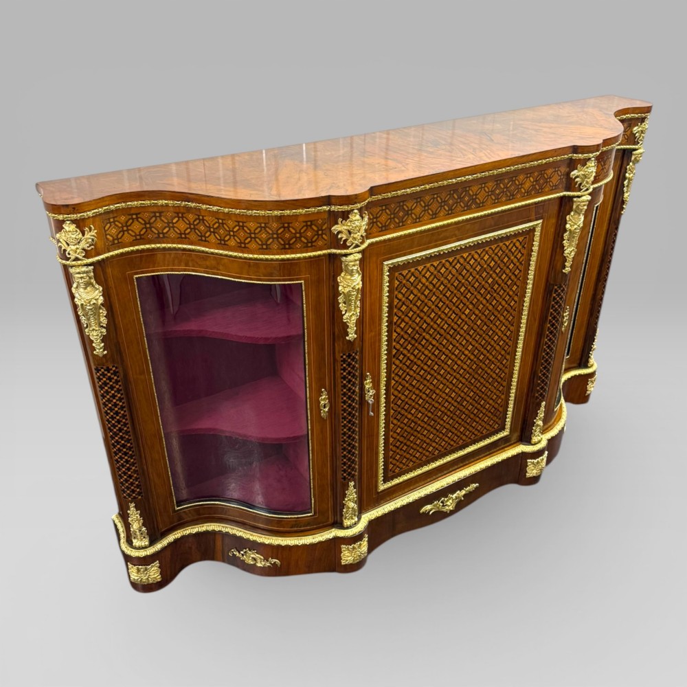 A Victorian walnut serpentine credenza featuring geometric marquetry, gilt ormolu mounts, a central panel door, and two curved glass side doors revealing lined display shelves. View IV