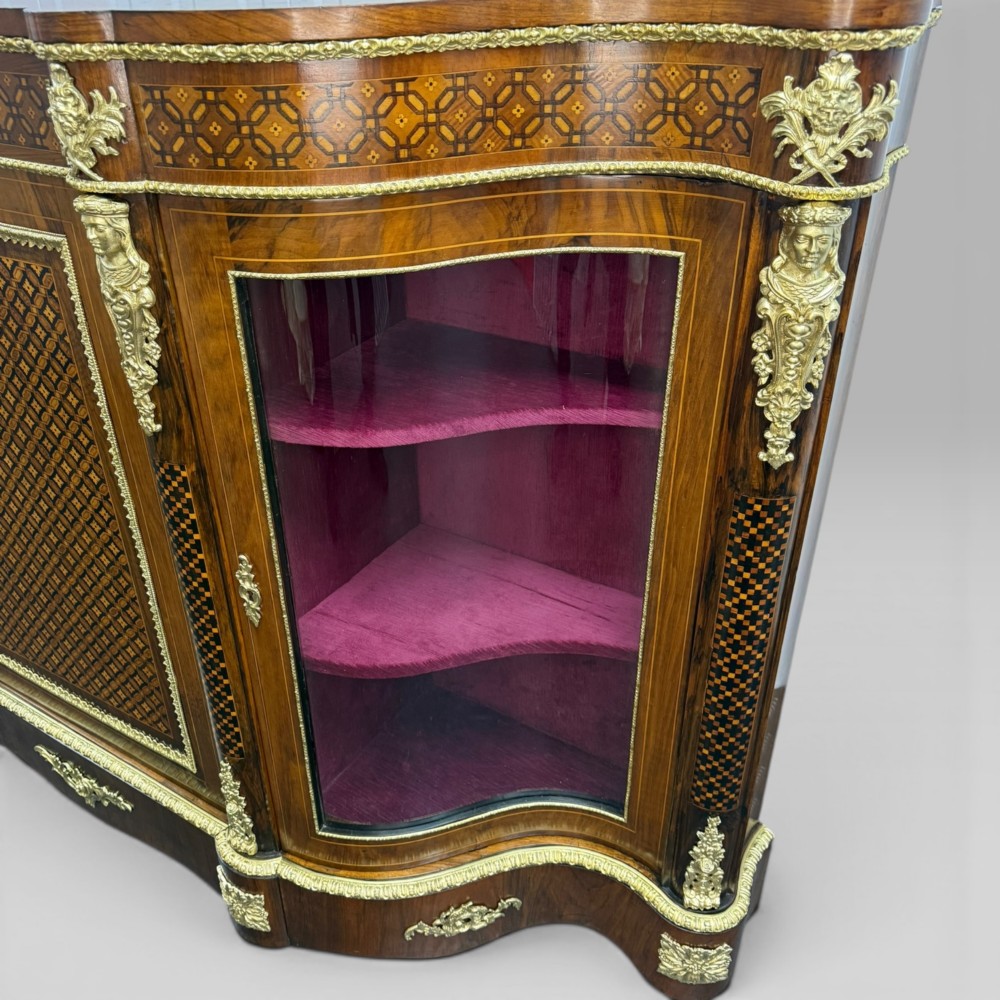 A Victorian walnut serpentine credenza featuring geometric marquetry, gilt ormolu mounts, a central panel door, and two curved glass side doors revealing lined display shelves. View VI