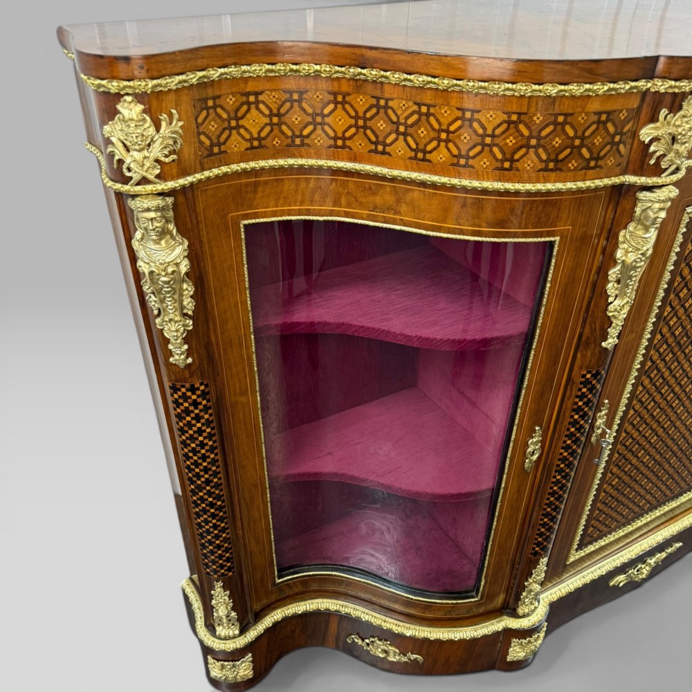 A Victorian walnut serpentine credenza featuring geometric marquetry, gilt ormolu mounts, a central panel door, and two curved glass side doors revealing lined display shelves. View VII