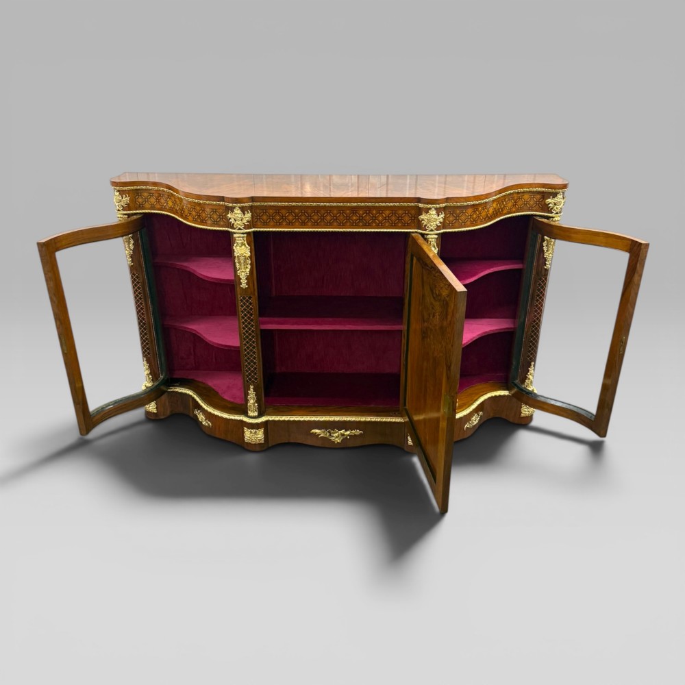 A Victorian walnut serpentine credenza featuring geometric marquetry, gilt ormolu mounts, a central panel door, and two curved glass side doors revealing lined display shelves. View IX