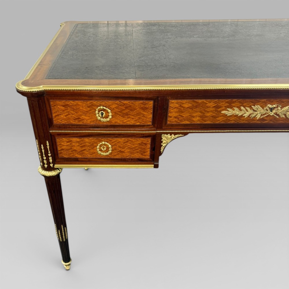 A Louis XVI style mahogany and parquetry writing desk featuring a dark leather top, gilt-bronze mounts, five drawers, and fluted tapered legs. View VI